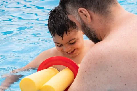 Disabled boy with a float laughs as he plays and swims in a pool with his father Stock Photos
