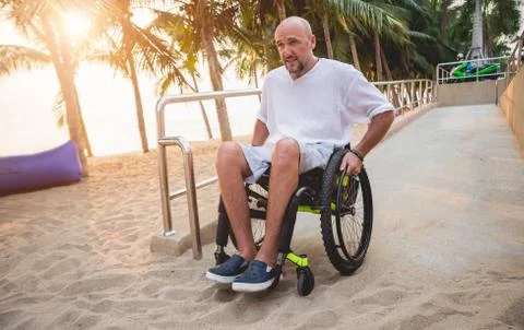 Disabled man in a wheelchair moves on a ramp to the beach. Stock Photos