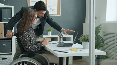 Disabled office worker in wheelchair talking to male colleague looking at laptop Stock Footage