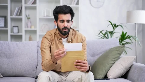 Disappointed young handsome man reading letter with bad news sitting on sofa at  Stock Footage 252203300