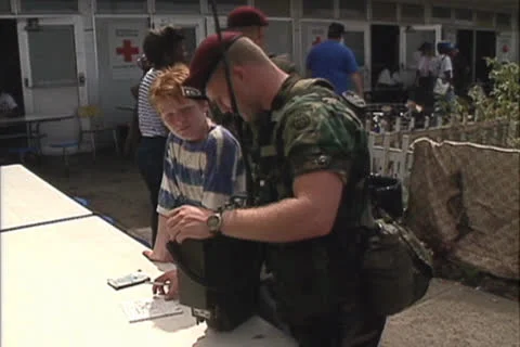 Disaster, US Airborne Trooper, talks to young boy pan right to food trucks Stock Footage 25828382