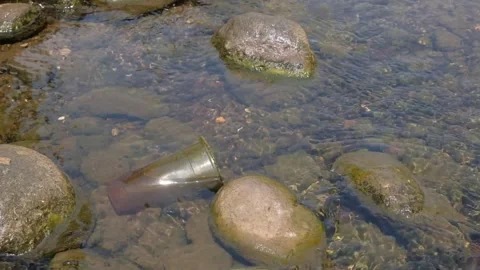 Discarded plastic cup submerged between rocks in shallow river water Video stock 347571728