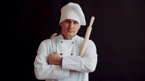 Disgruntled angry chef with rolling pin in white kitchen suit on black wall Vidéo 95746364