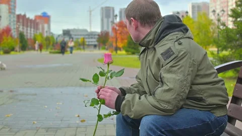 Disgruntled, angry guy sits on a park bench with a rose flower in his hand Stock Footage 165188418