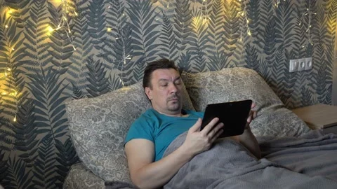 A disgruntled middle-aged man reads the news on a tablet while lying on his 스톡 동영상 148447947