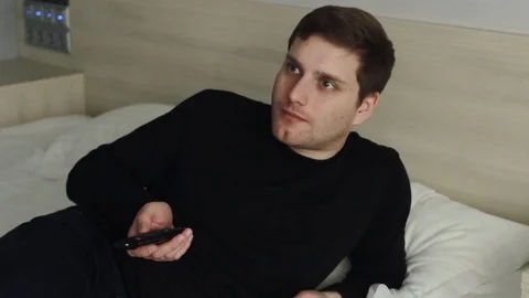Dissatisfied young man with TV remote watching tv on bed Stock Footage