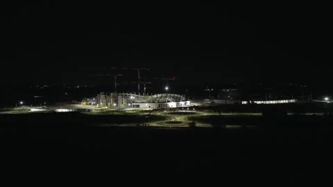 Distant construction cranes tower above illuminated oval stadium framework at Stock Footage 346786141