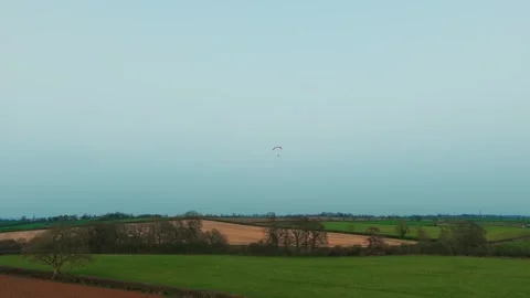 Distant paramotor pilot gliding over patchwork farmland fields forest edges and Stock-Footage 335683429