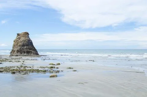 Distant rock stack aligned with gentle surf and expansive clouded sky on a .. Stock Photos