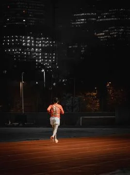 Distant runner on a red running track with lit buildings at night Фото