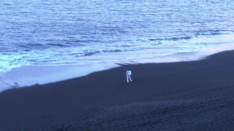 Distant silhouette of man in white running along coastline of Reynisfjara beach Stock Footage 257790849