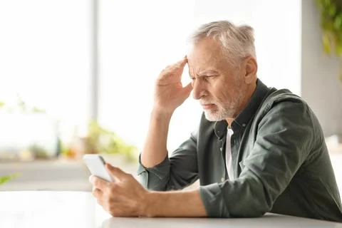 Distressed elderly man suffering headache while looking at smartphone at home 库存照片