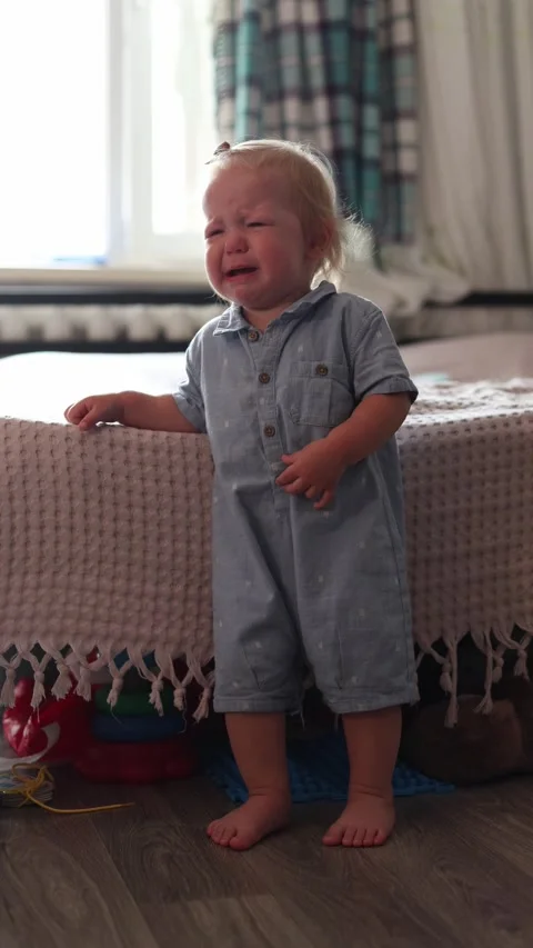 A distressed toddler crying while standing beside a bed in a bright and cozy Stock Footage 345610999