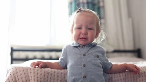 A distressed toddler standing by a bed in a bright bedroom during the daytime at Stock Footage 345610994