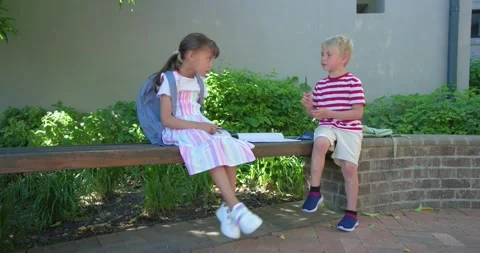 Diverse children sitting on bench in courtyard after girl glancing, boy flipping Stock Footage 336957760