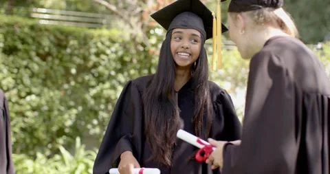 Diverse classmates in gowns pulling smartphone under tree and beaming for Stock Footage 316476367