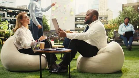 Diverse colleagues discuss planning notes beside laptop at outdoor discussion Stock Photos