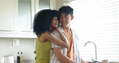 Diverse couple adjusting window blinds, embracing at kitchen countertop, sharing Stock Footage 342719216