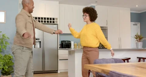 Diverse couple clasping hands by sofa, dancing from living room into kitchen, Stock Footage 344316601