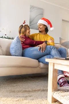 Diverse couple cuddling on light-colored sofa in living room, wearing Santa hat Stock Photos