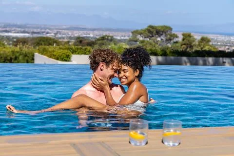 Diverse couple embracing in infinity pool at rooftop wooden deck in swimwear Foto stock
