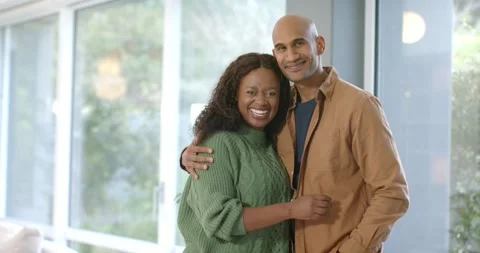 Diverse couple exchanging smiles at large glass window, draping arms and sharing 스톡 동영상 317965133
