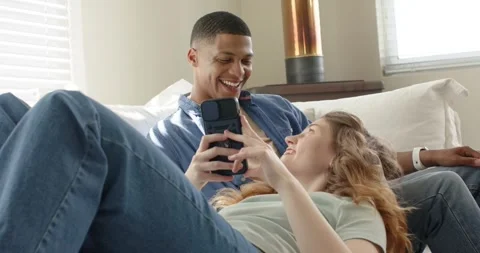 Diverse couple grabbing smartphone tapping screen scrolling images on sofa at Stock Footage 342018778