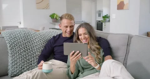 Diverse couple on gray couch reacting to tablet content while woman tapping and Stockbeeldmateriaal 340965440