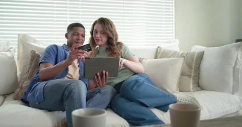 Diverse couple holding tablet and tapping checkout to complete purchase on sofa Stock Footage 345071370