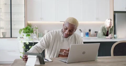 Diverse couple in kitchen, man using tablet then writing notes while she Stock Footage 344678737
