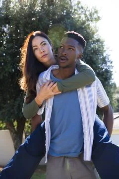 Diverse couple laughing while man giving piggyback ride in backyard near tree, Stock Photos