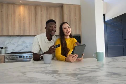 Diverse couple leaning and using tablet at kitchen island, holding credit card, Photos