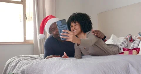 Diverse couple lying on bed reaching smartphone, framing selfie with Santa hat Stock Footage 335514085