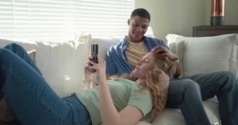 Diverse couple lying on white sofa woman scrolling flip phone sharing clip man Vídeos de archivo 341089906