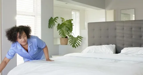 Diverse couple in pajamas making bed, placing pillows in sunlit master bedroom Stock Footage 341931425