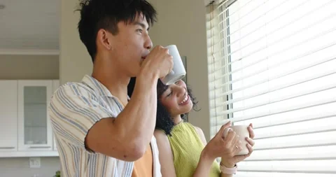 Diverse couple peering at kitchen window behind blinds holding mugs, leaning and Stock Footage 344948276