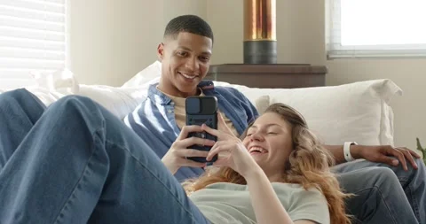 Diverse couple picking up smartphone on couch in living room, loading funny Stock Footage 344326011