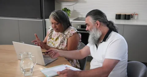 Diverse couple pointing at charts in kitchen while reviewing data on laptop and Stock Footage 337536843