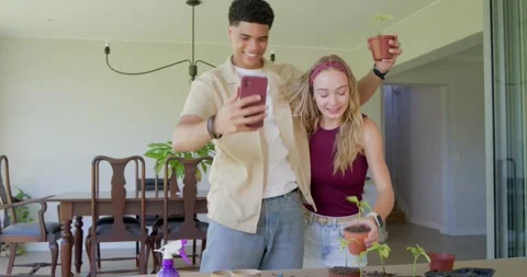 Diverse couple posing for phone then potting seedlings at table with soil and 库存影片 340974598