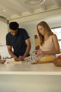 Diverse couple preparing meal at kitchen island, chopping herbs and cracking egg 스톡 사진