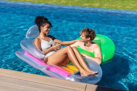Diverse couple relaxing in backyard pool on clear lounge and green tube, woman Foto stock