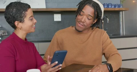 Diverse couple scrolling smartphone and leaning over laptop in kitchen sharing Vídeo Stock 336535452