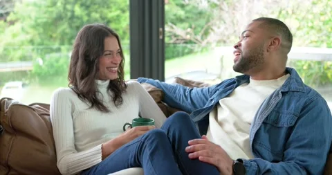 Diverse couple settling onto leather sofa by window, talking and laughing over Vídeo Stock 344330802
