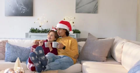 Diverse couple settling on sofa at home wearing hats sipping mugs sharing cookie Stock Footage 344226245