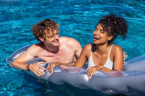 Diverse couple sharing clear float leaning, laughing in blue pool, sunglasses Fotos Stock