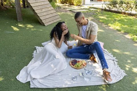 Diverse couple sharing fruit on blanket in garden, woman in white dress man Fotos Stock