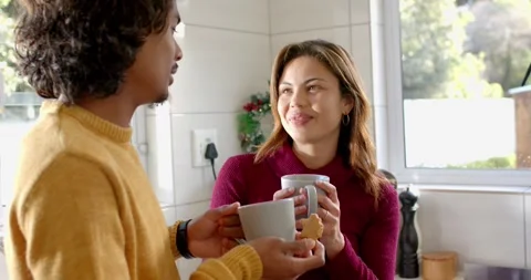 Diverse couple sharing gestures conversing in kitchen by window holding mugs and Stock Footage 344226062