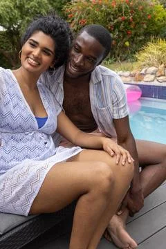 Diverse couple sitting at backyard pool, woman in white lace cover-up, pink Fotos de archivo