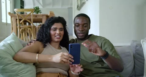 Diverse couple sitting on couch at home, woman holding blue phone, man pointing, Stock Footage 340981495