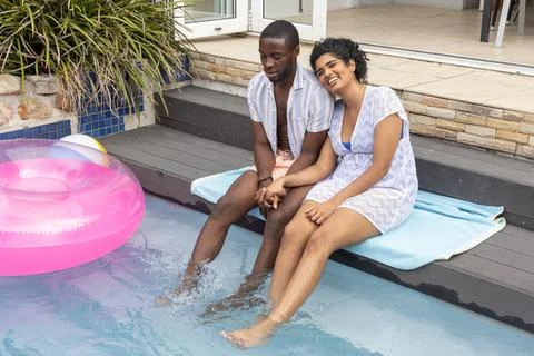 Diverse couple sitting on deck steps at backyard pool wearing swimwear holding Stock Photos